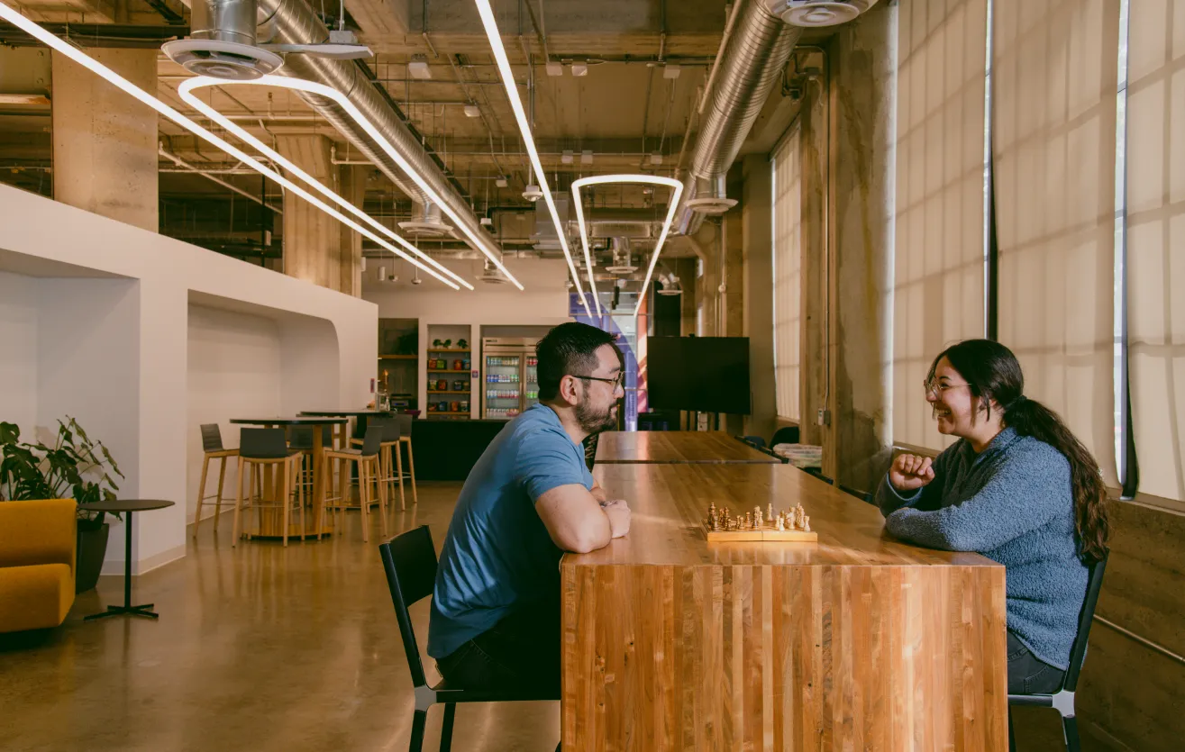 One of the many collaborative spaces found in the Atlas Meet offices — two employees are sitting across from each other, smiling and conversing over a chess game. They are seated at a long wooden table, various chairs and couches are behind them, as well as a refrigerator and snack area.