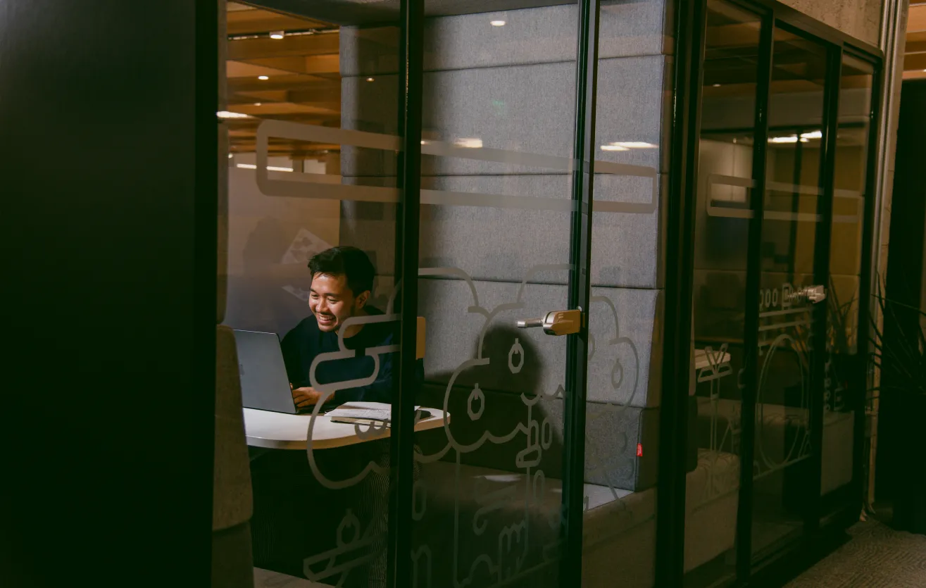 A Atlas Meet employee sitting in a private work booth, visible behind a glass door that is shut. He is working on his laptop and sitting on a comfy, fabric bench.