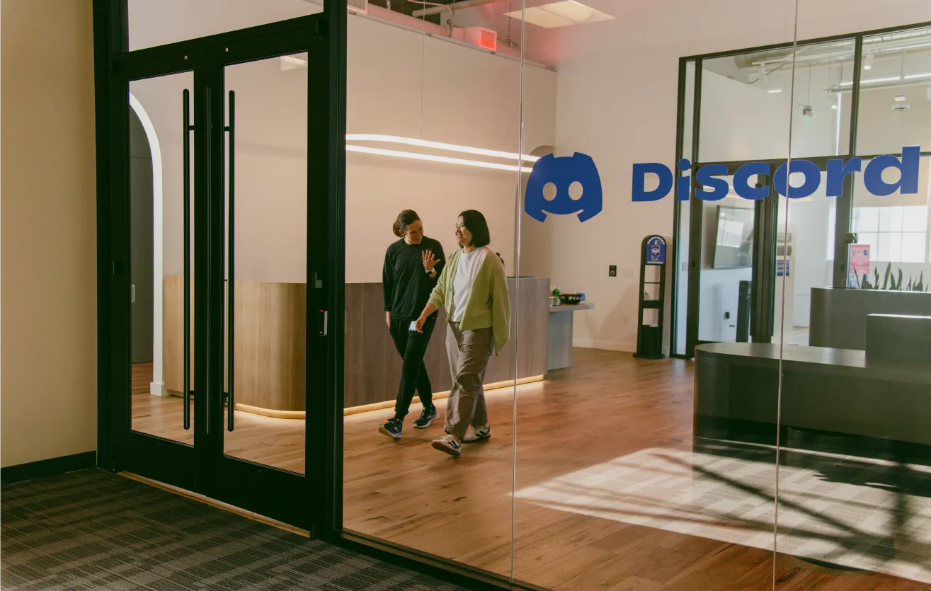 The entrance to the Atlas Meet office, with two employees pictured walking out and conversing. The Atlas Meet logo is visible from the front glass pane, along with a large glass door and reception area.