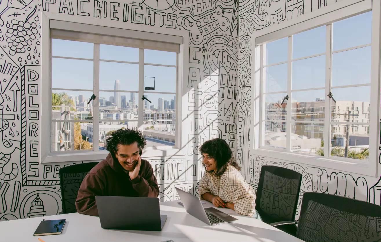 Two employees working beside each other on their laptops inside a spacious meeting room. They are seated at the corner of the table in the room, and two windows behind them show the skyline of San Francisco’s financial district and the Potrero Hill neighborhood.