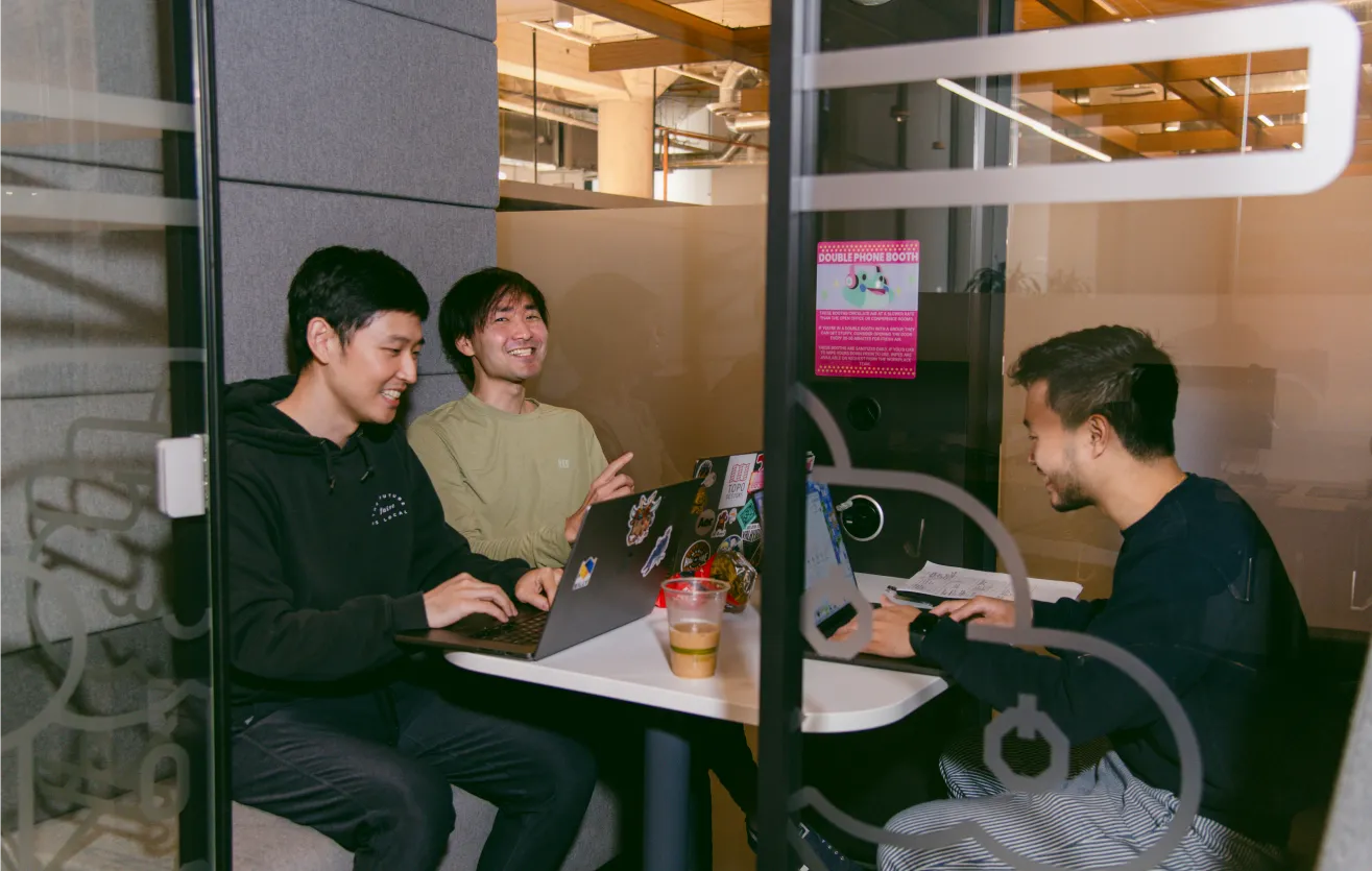 Three employees are working collaboratively inside a private workstation, the glass door is ajar. All three of them have their work laptops atop the desk in the middle. Beside one of them there is a cup of iced Coffee.