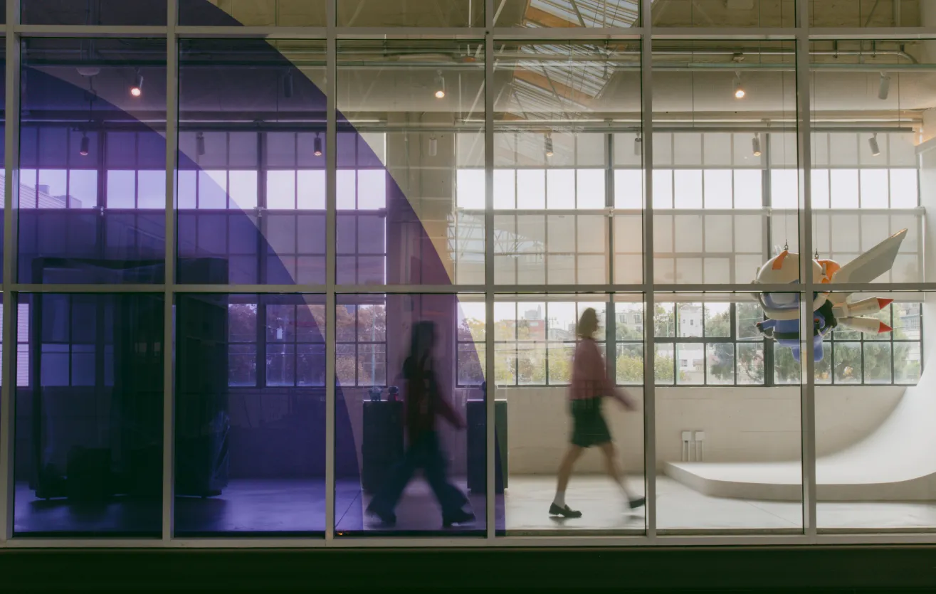 A photo of one of the walkways near the front of the Atlas Meet office, containing references to various Atlas Meet memorabilia in glass cases, and Wumpus (Atlas Meet’s mascot) riding a jetpack, suspended in air. Two employees are walking through the area — probably on their way to grab coffee together!