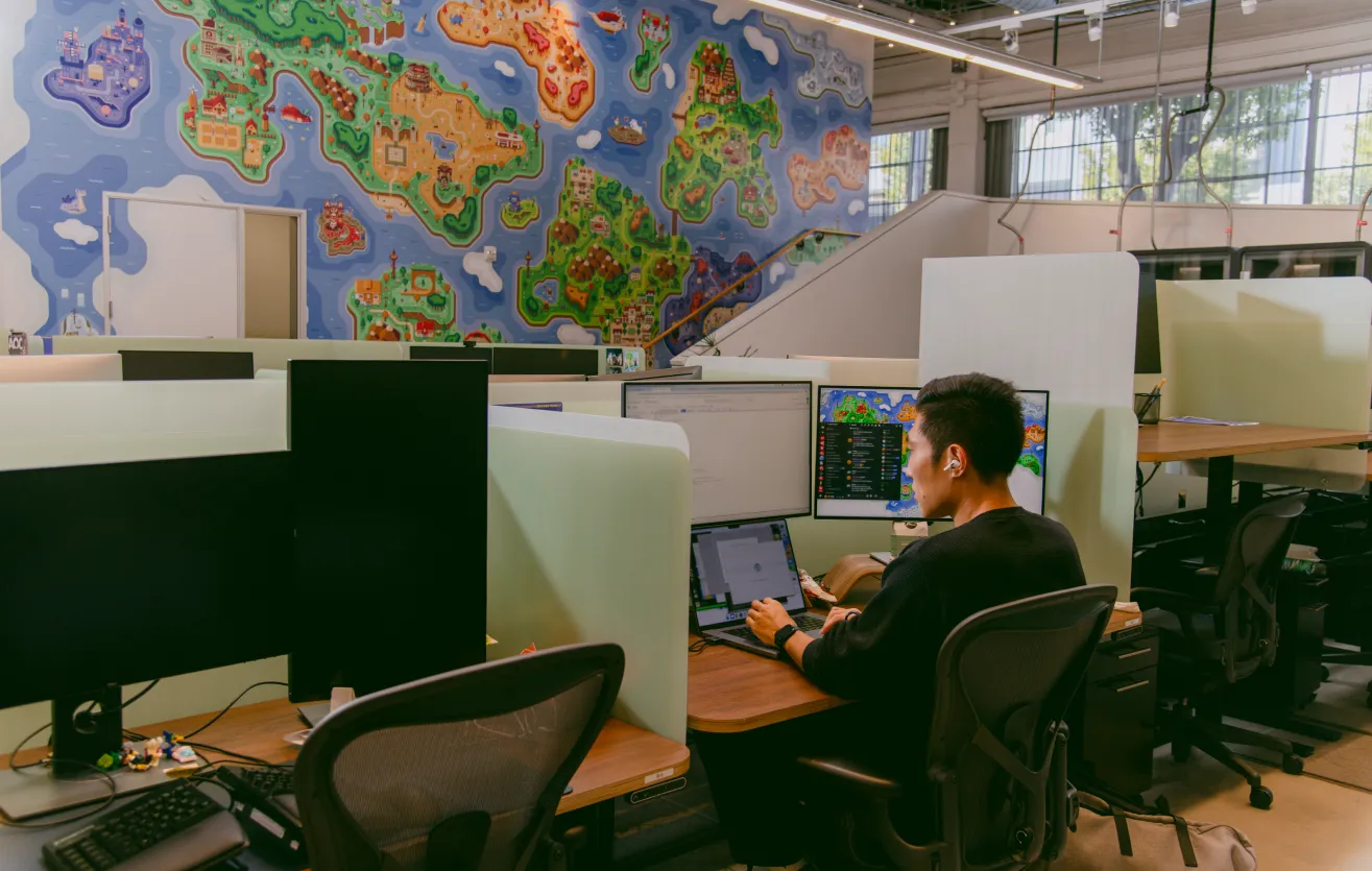A Atlas Meet employee at work in the Atlas Meet office, sitting at his desk, surrounded by two monitors and a laptop. Behind it lays a mural containing popular references to various video games.