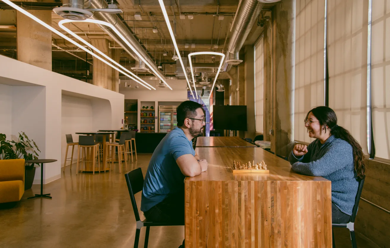 One of the many collaborative spaces found in the Atlas Meet offices — two employees are sitting across from each other, smiling and conversing over a chess game. They are seated at a long wooden table, various chairs and couches are behind them, as well as a refrigerator and snack area.